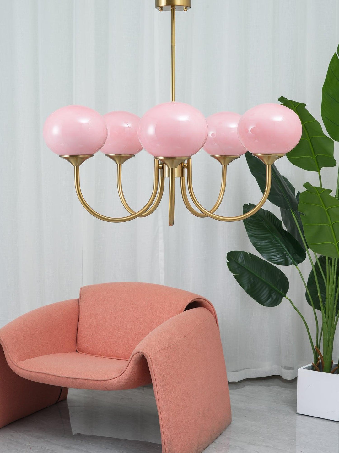 Rosa Kronleuchter mit goldenen Armen, modernes Design, über rosa Sessel, vor weißen Vorhängen.