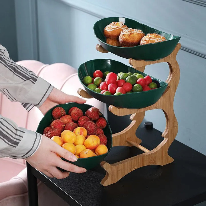 Dreistufiger Obstständer aus Holz mit grünen Schalen, gefüllt mit Obst und Gebäck.