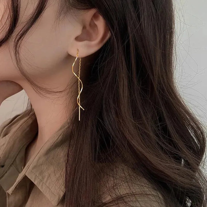 Frau mit langen, goldenen Ohrringen, welliges Haar, braune Bluse, eleganter Schmuck.