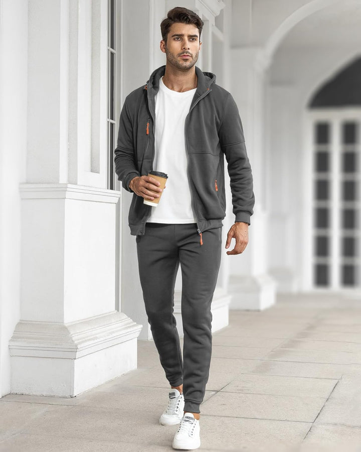 Mann in grauem Jogginganzug mit Kapuze, hält Kaffeebecher, weiße Sneaker, urbaner Hintergrund.