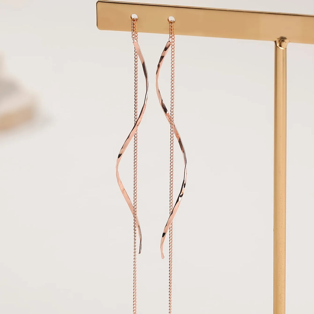 Roségoldene, lange Ohrringe mit geschwungenem Design, hängend an einem goldenen Ständer.
